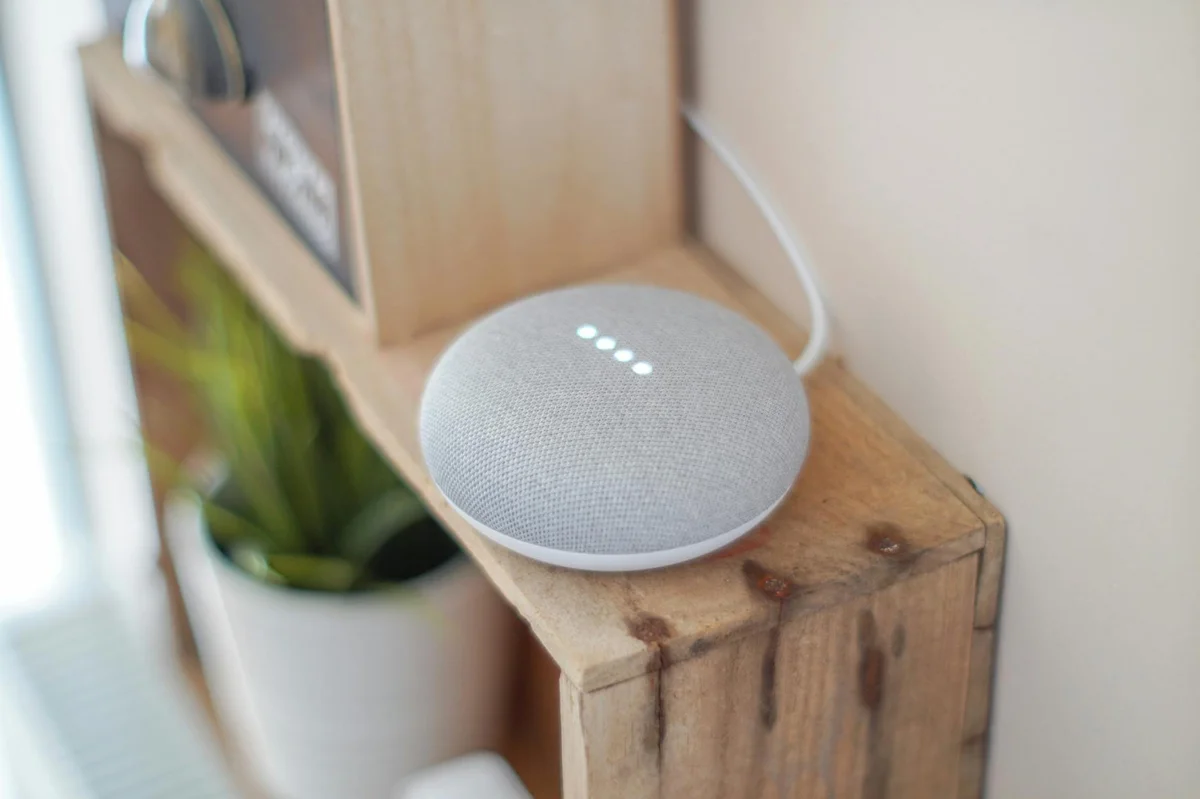 Tareas programadas y agentes IA automáticos: trabajo mientra... — A Google Home Mini smart speaker on a wooden shelf, blending technology with hom