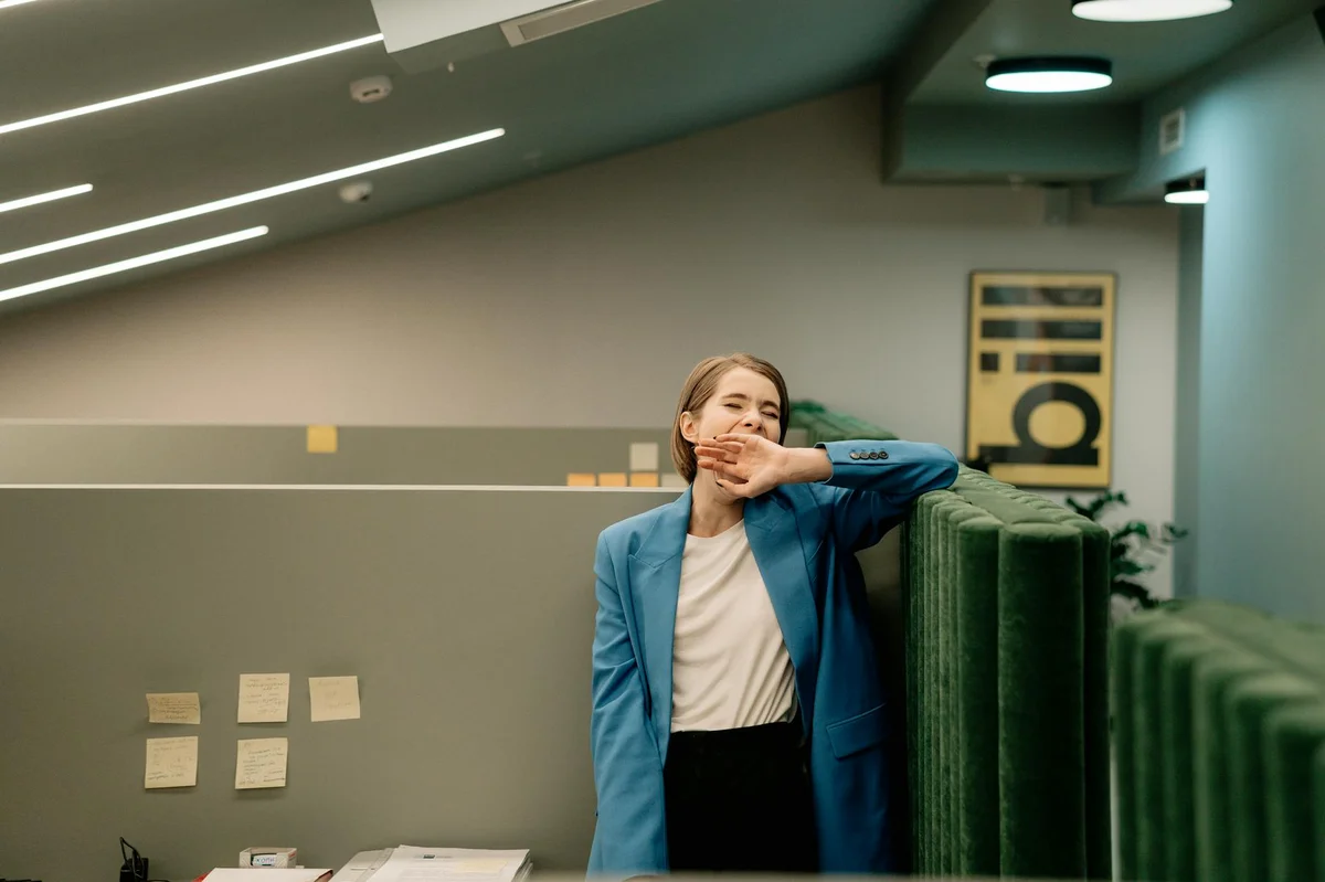 Dejar de apagar incendios: el sistema para trabajar con calm... — Exhausted woman in blue blazer yawning during workday in corporate office.