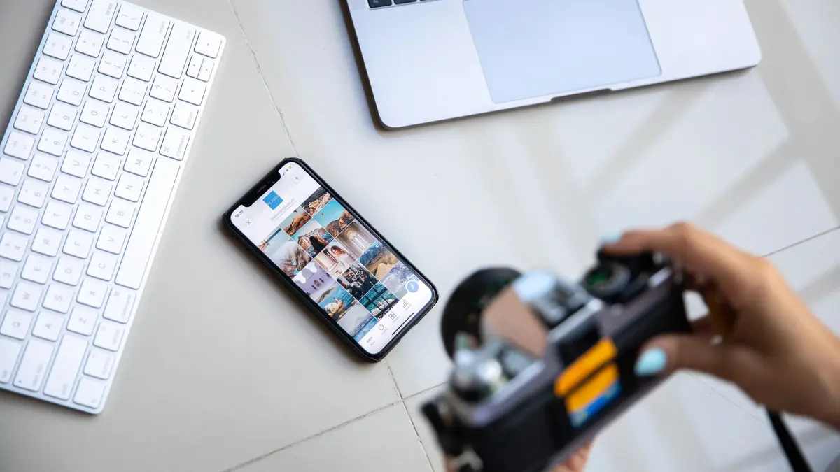Person using vintage camera with smartphone and laptop in creative setup.