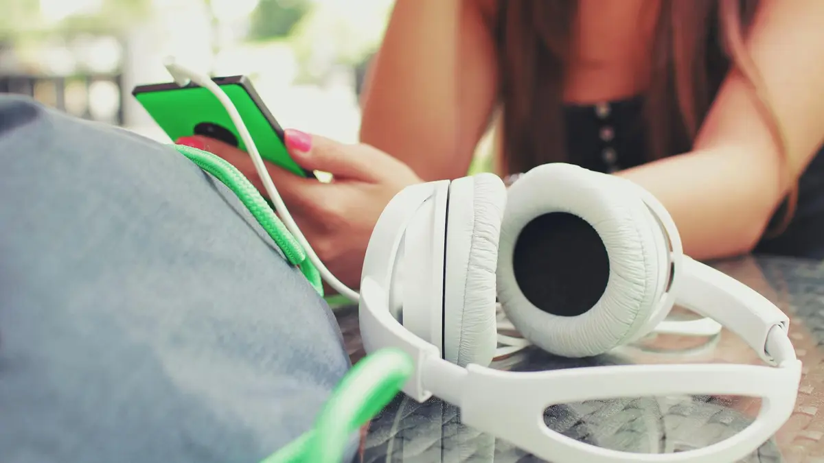 A woman listens to music with headphones, using a smartphone at an outdoor café.
