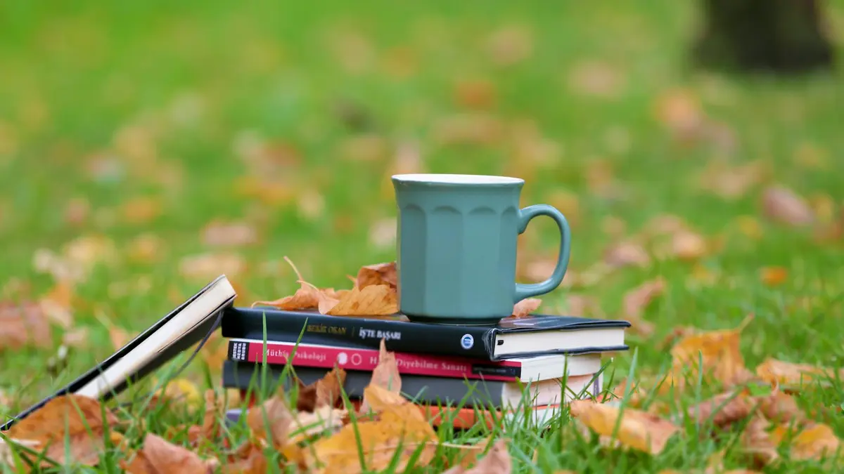 A serene autumn setup of books and a cup on grassy field with fallen leaves.