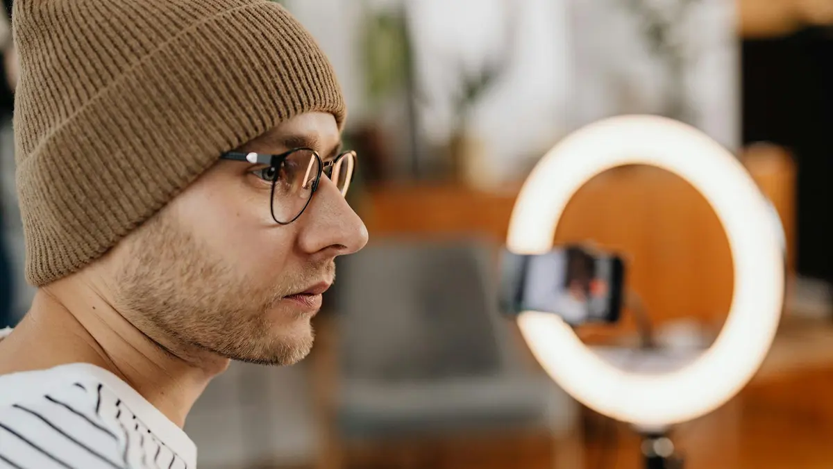 A man wearing eyeglasses and a knitted cap records a video at home using a smartphone and ring light