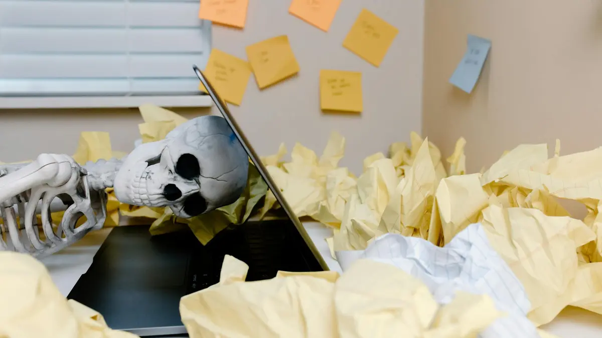 A humorous skeleton slumped over a laptop among crumpled papers in an office setting.