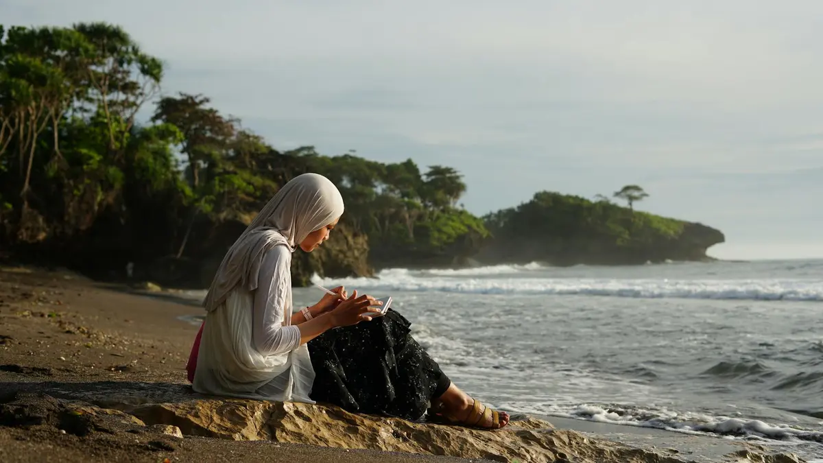 A woman in a hijab reads by the beach in Jawa Barat, Indonesia during a serene day.
