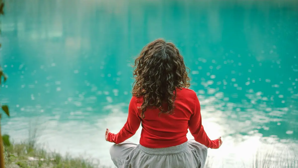 A woman in red meditates by a tranquil blue lake, reflecting calm and serenity.