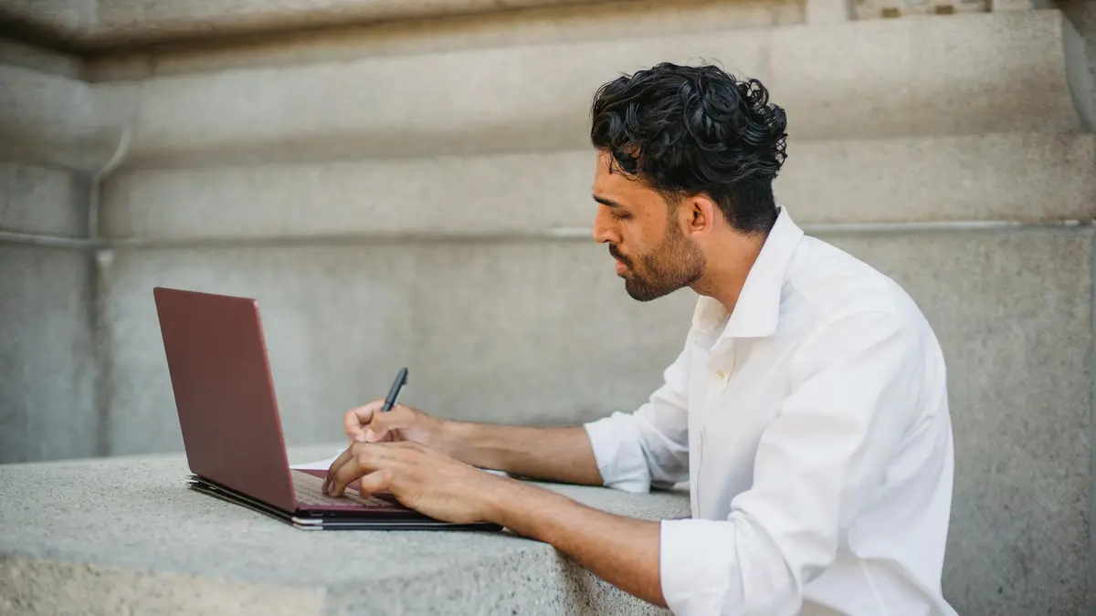 A man writing on a laptop outdoors, seated on a stone surface and focused on work.